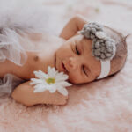 Newborn lying on pink fluffy blanket, holding a daisy. She has grey and with tuille over her tummy and a white and grey headband on.