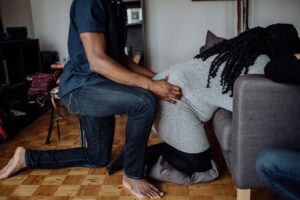 Pregnant Woman kneeling over chair while birth partner kneels behind her massaging her lower back